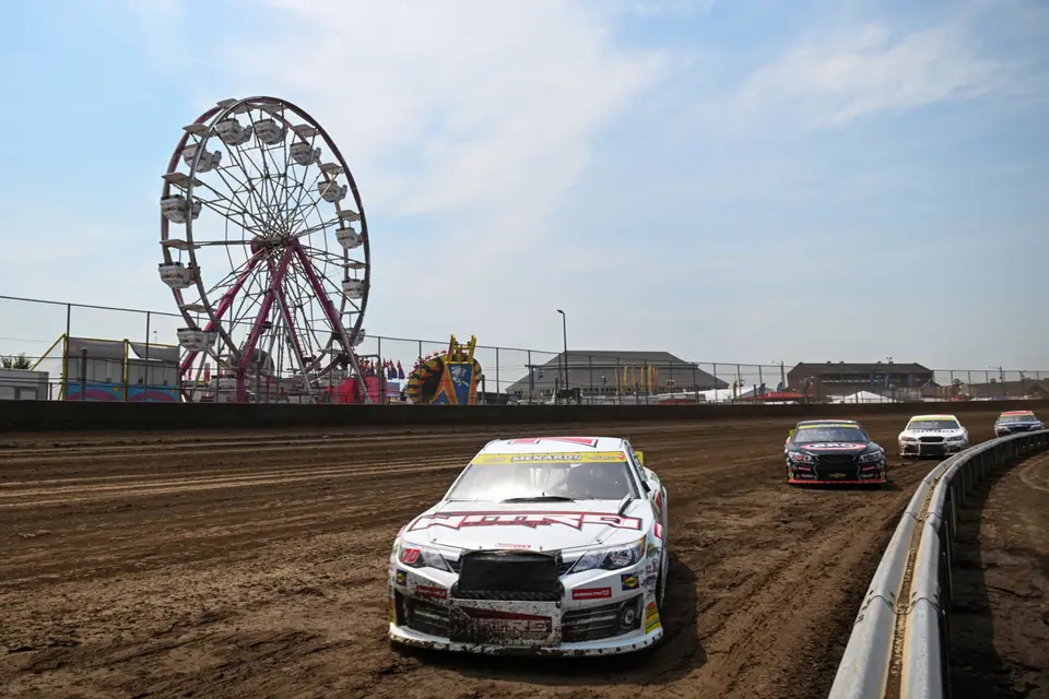 Dirt race car passing a Ferris wheel at an evening event