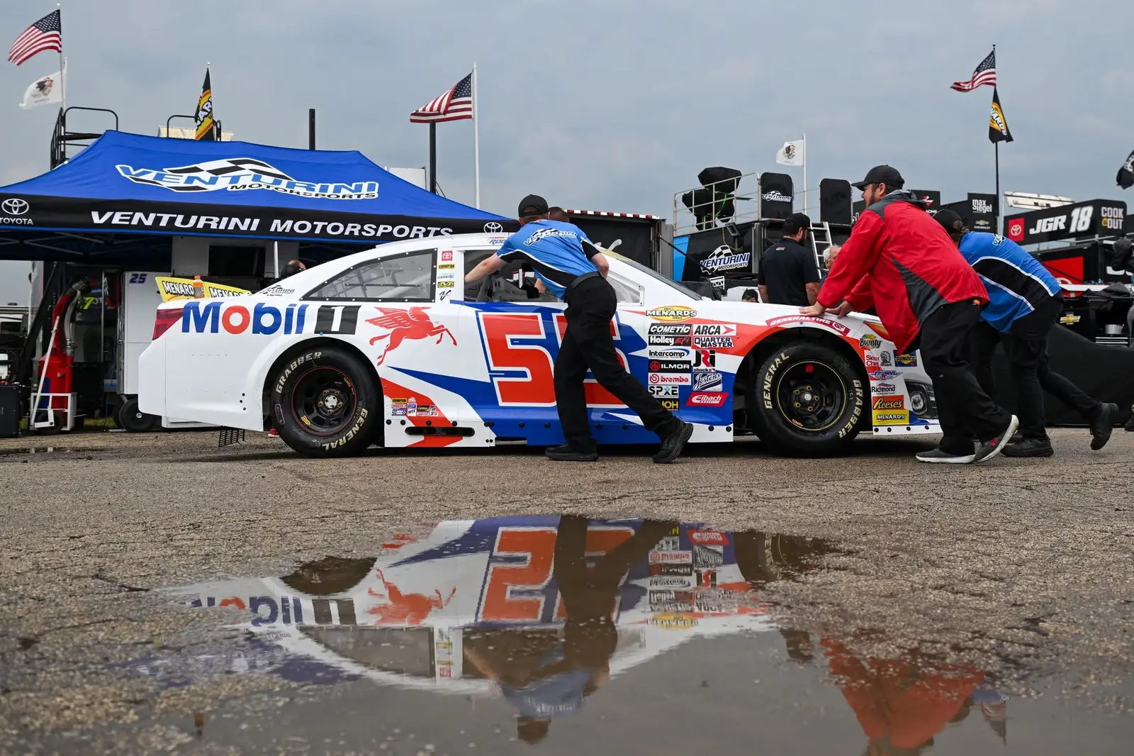 ARCA crew pushing a race car through the paddock above a rain puddle reflection