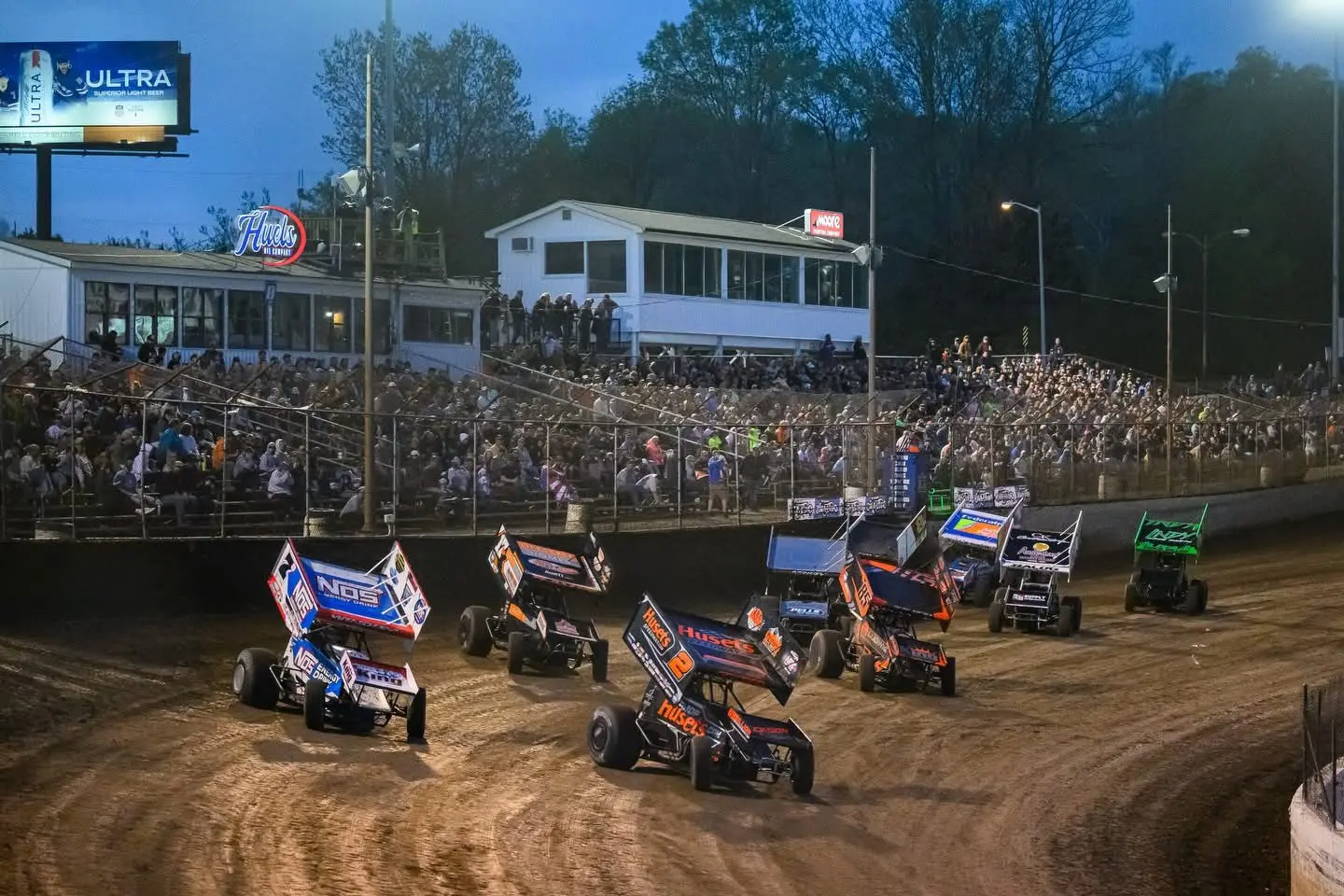 Winged sprint cars entering a dirt-track corner in front of a packed grandstand